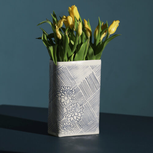 A large ceramic vase filled with yellow tullips resting on a dark table. The vase has a geometric pattern in blue