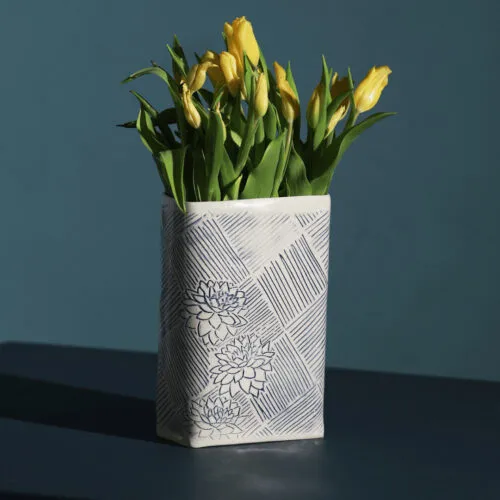 A large ceramic vase filled with yellow tullips resting on a dark table. The vase has a geometric pattern in blue